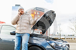 A young man looks angry and frustrated near his broken car and speaks on his mobile phone,