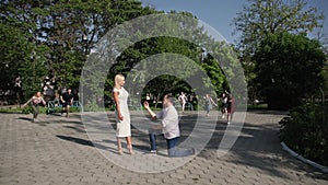 young man in love is on his knees and makes an offer with help of ring to his beloved woman, crowd of people runs to