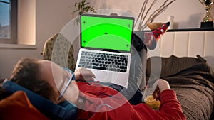 Young man lying on the bed with laptop and eating cheeps - green screen