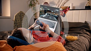 Young man lying on the bed playing race video game on the laptop with gamepad