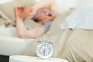 Young man lying in bed yawning near alarm clock