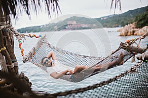 Young man lying in a hammock by the ocean. A guy is resting in a hammock against the backdrop of the mountains by the sea