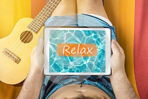 Young man lying on a hammock with tablet device looking at water background with word Relax written on it