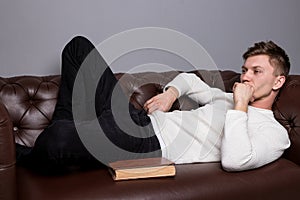 Young man is is lying on a leather sofa and reading a book