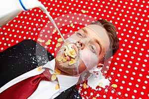 Young man lying on red vintage table cloth with corn flakes in mouth. Milk pouring inside mouth. Breakfast time. Food