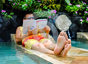 Young man lying in swimming pool and reading magazine