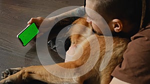 Young man lying on wooden floor with dog uses smartphone to watch movie or video. Green screen, horizontal mockup. Owner