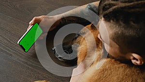 Young man lying on wooden floor with dog uses smartphone to watch movie or video. Green screen, horizontal mockup. Owner