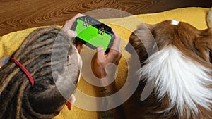 Young man lying on yellow blanket on bed with dog using smartphone with joystick for online game. Green screen