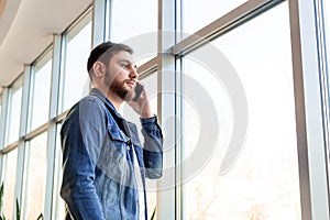 Young man making business call talking by phone indoors near window wall in casual jean jacket. Smart man in modern city office