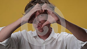 Young man making heart shaped hands gesture on isolated on yellow background. The winking face of a handsome young man