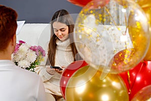Young man making surprise for his girlfriend holding flowers in his hands and colored balloons while she sitting on a