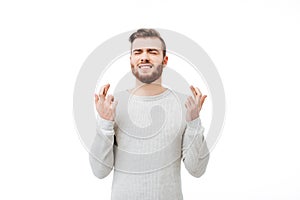 Young man making a wish emotionally with finger crossed gesture over white background. Guy praying with eyes closed
