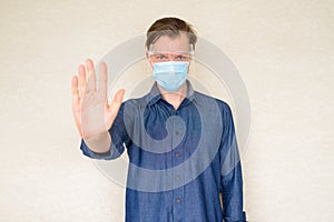 Young man with mask and face shield showing stop gesture against concrete wall