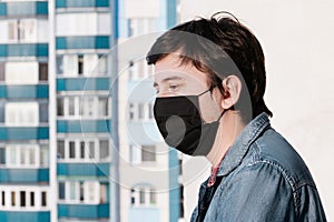 A young man in a medical mask black looks into the distance against the background of city buildings