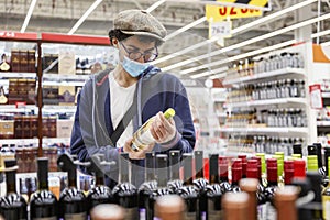 A young man in a medical mask, glasses and a cap chooses alcohol in the store. Depression and holidays during the coronavirus