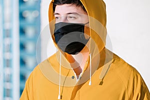 A young man in a medical mask, in a jacket yellow looks ahead against the blurry background of the city