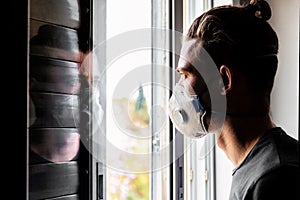 Young man in a medical mask, looks out the window and is reflected in the glass
