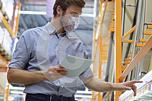 Young man in medical mask shopping or working in a hardware warehouse standing checking supplies on his tablet