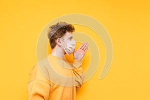 Young man in a medical mask shouts to the side with his hand to his face on a yellow background. Guy in the protective white mask