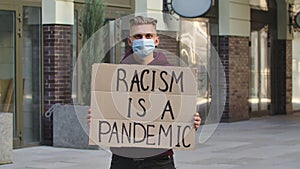 Young man in medical mask stands with a cardboard poster RACISM IS A PANDEMIC in a public place outdoor. No racism