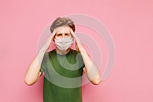A young man in a medical mask suffers from a migraine on a pink background, touches his head with his hands and looks wearily into