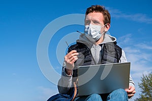 Young man in medical mask works or studies remotely during pandemic on Sunny day sitting on grass in park outdoors. Spring relax