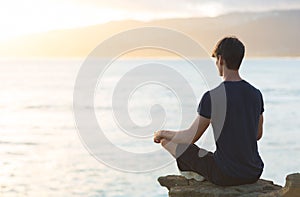 Young man meditating on top ocean cliff during sunset.