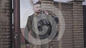 The young man in military closes with big bag entering the gate. A man returns from the military service home. Leaving