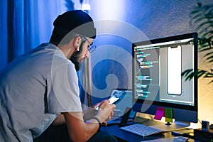 Young man mobile developer writes program code on a computer and check app in smartphone, programmer work in home office