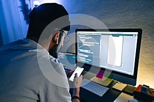 Young man mobile developer writes program code on a computer and check app in smartphone, programmer work in home office