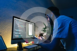 Young man mobile developer writes program code on a computer and check app in smartphone, programmer work in home office