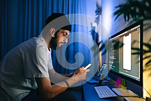 Young man mobile developer writes program code on a computer and check app in smartphone, programmer work in home office