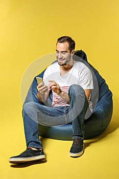 Young man with mobile phone sitting on beanbag chair against color background