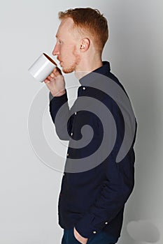 A young man with a mug of tea or coffee. He pleased. White background. Redhead male with white mug.