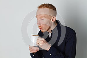 A young man with a mug of tea or coffee. He pleased. White background. Redhead male with white mug.