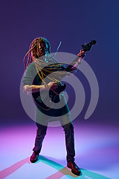 Young man, musician, expressive guitarist with dreadlocks playing guitar, performing solo against dark purple background
