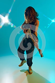 Young man, musician with long hair emotionally and energetically playing guitar in stage against blue, cyan background