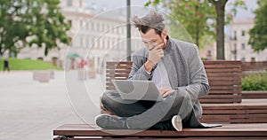 Young man with mustaches and a beard sitting cross-legged on wooden bench in the square, typing on the laptop keyboard