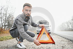 A young man near a broken car with an open hood on the roadside.