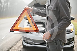 A young man near a broken car with an open hood on the roadside.