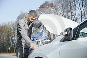 A young man near a broken car with an open hood on the roadside.