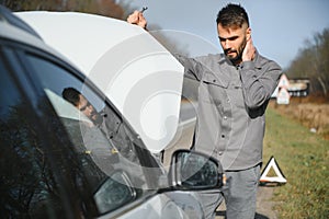 A young man near a broken car with an open hood on the roadside.