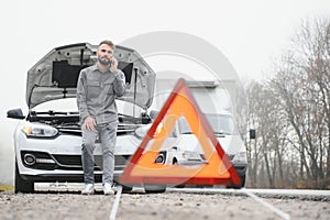 A young man near a broken car with an open hood on the roadside.