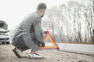 A young man near a broken car with an open hood on the roadside.