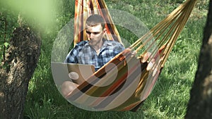 Young man opening laptop computer and starting to work while lying in hammock outdoors. Man working on notebook at green garden. F