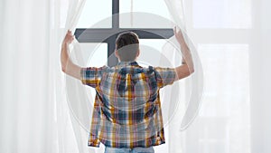 Young man opening window curtain at home