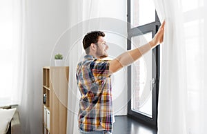 Young man opening window curtain at home