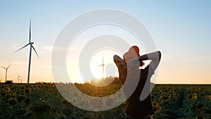 Young man in orange cap in yellow sunflowers crops field with wind turbines farm on sunset landscape.