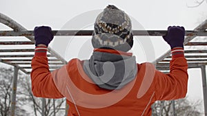 Young man in orange jacket doing pullups on horizontal bars outdoors in winter